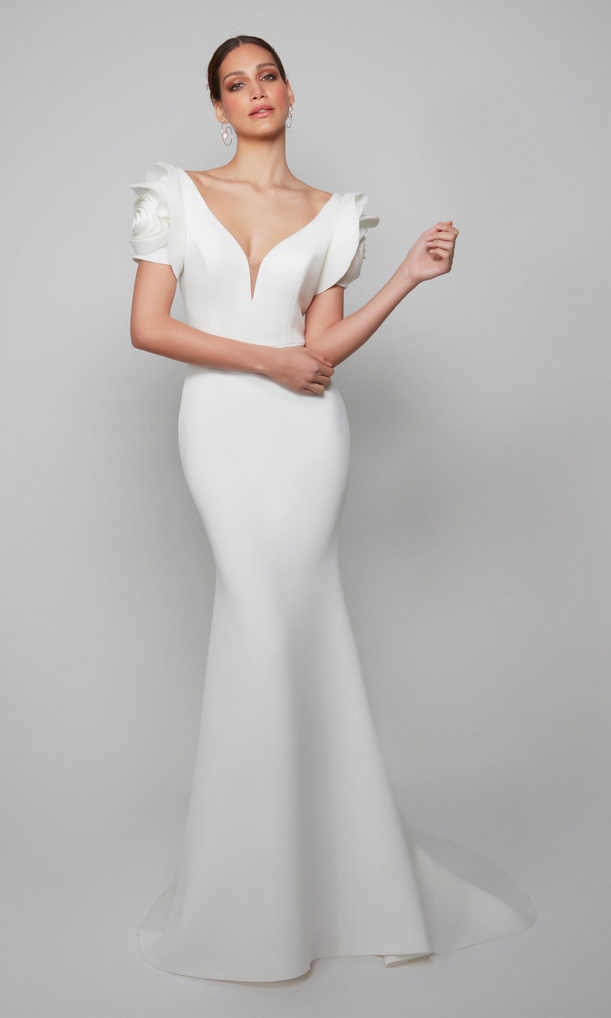 A woman in the Alyce 7066 long formal dress—a fitted white gown with a deep V neckline, sculpted short sleeves, and a mermaid silhouette—stands before a plain light background.